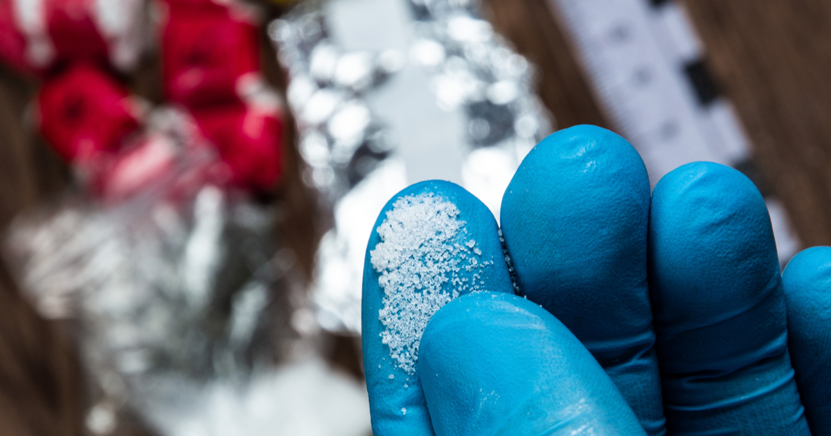 Blue-gloved fingers holding granular white substance, blurred objects in background.