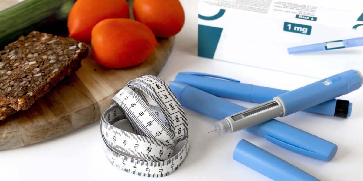 Bread, tomatoes, tape measure, and blue pen-like medical devices on a wooden board.