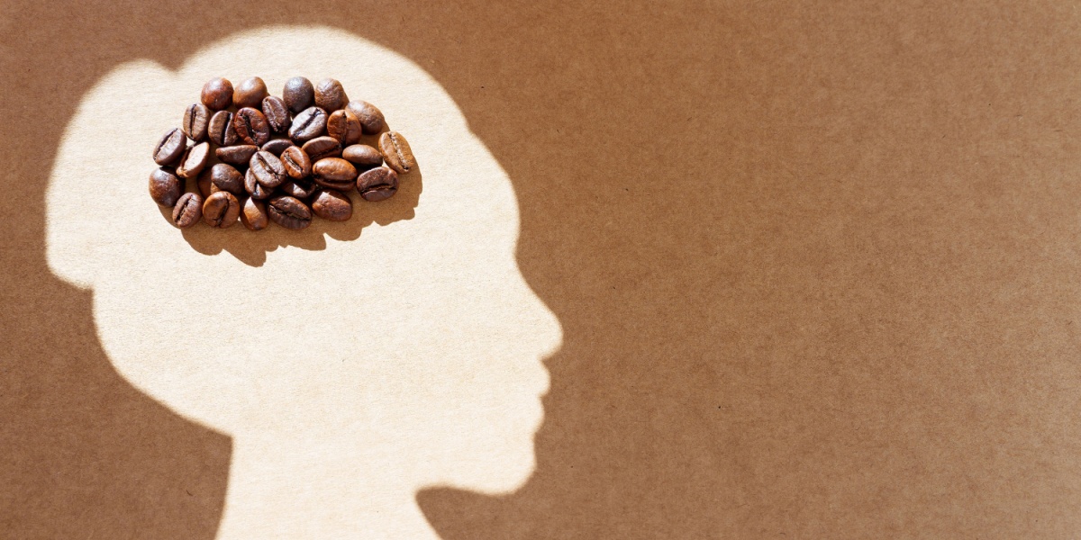 Pile of roasted coffee beans arranged into a brain shape inside a light female head silhouette on a craft paper background.