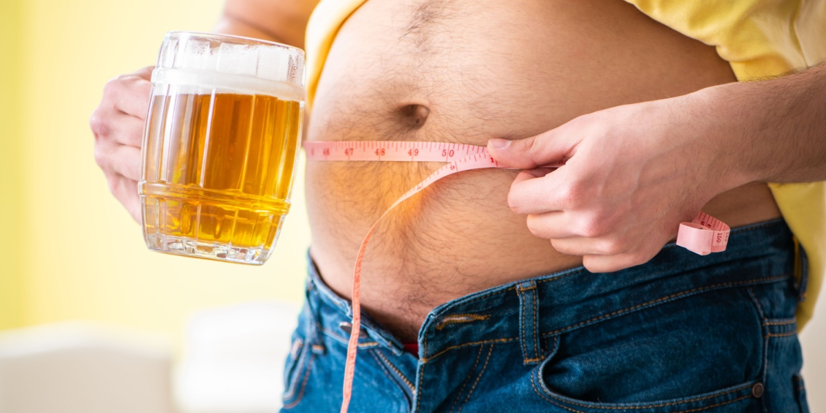 a close up photo of a man's exposed bloated belly while he's holding a mug of alcohol in one hand and a tape measure on the other