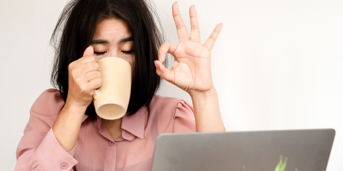 A half body shot of a woman drinking coffee in front of their work laptop, obviously disheveled and tired but still holding up an OK sign with her fingers