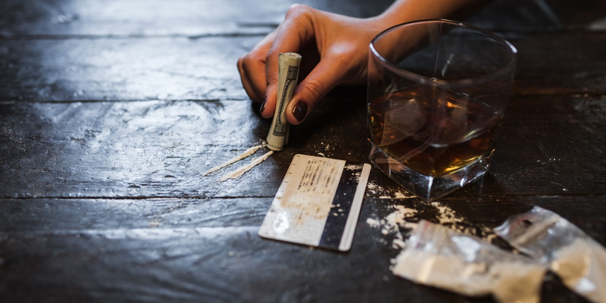 a photo of a manicured hand on a table holding up a rolled up dollar bill, there's also powdered cocaine and alcohol on the table