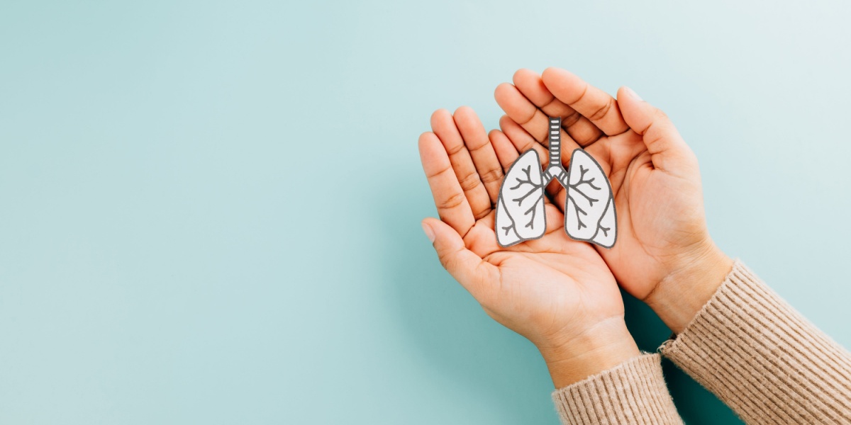 a top view of two hands gently cradling a cutout drawing of a pair of lungs over a soft teal background