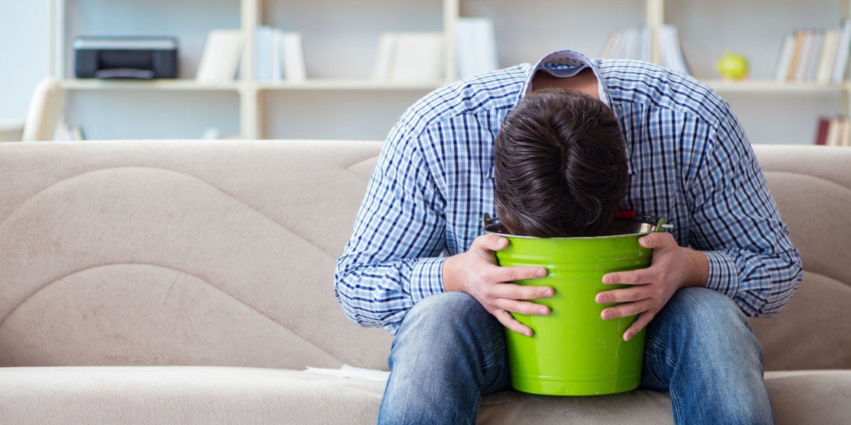 a photo of a man sitting in a sofa and hunched over a bucket due to weed hangover