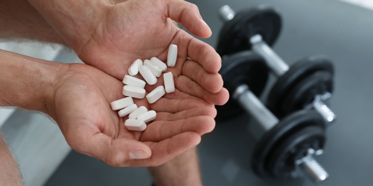 a close-up photo of a hand holding up Anabolic Steroid pills with a pair of dumbbells in the background