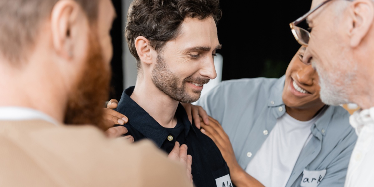 a close up of a Caucasian man being surrounded by his support group and being congratulated for his cocaine addiction recovery
