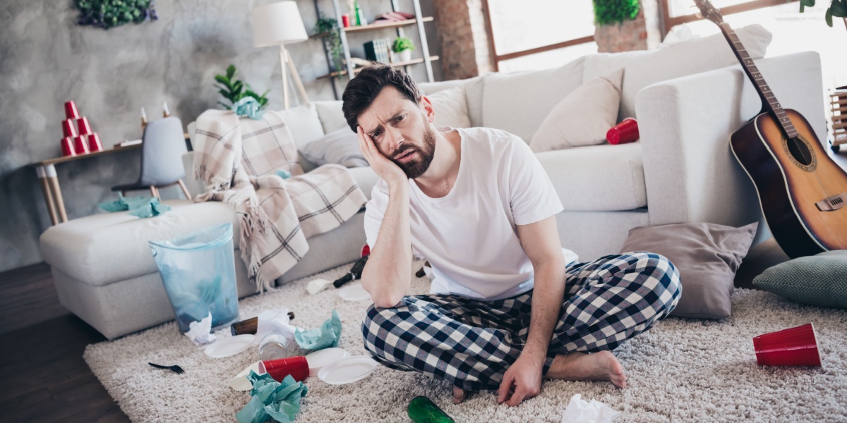 a Caucasian man sitting in the middle of a living room surrounded by cups, unable to remember anything from the previous night due to alcohol