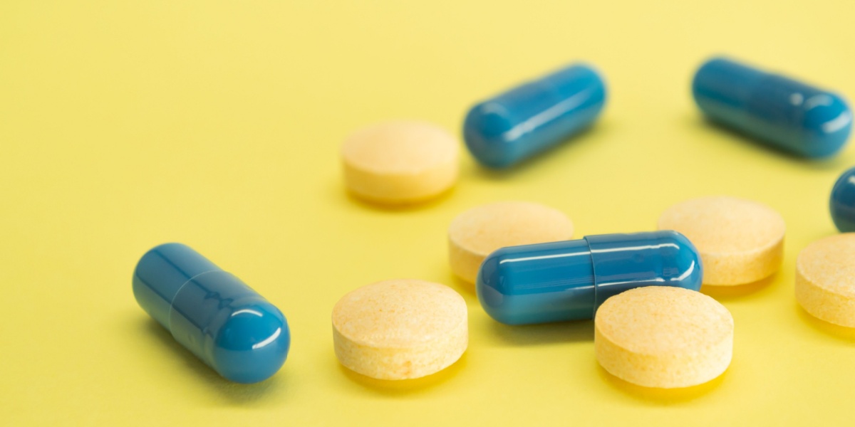 A close up photo of blue capsules and yellow circular pills scattered across a light yellow background, comparing dexedrine and Adderall