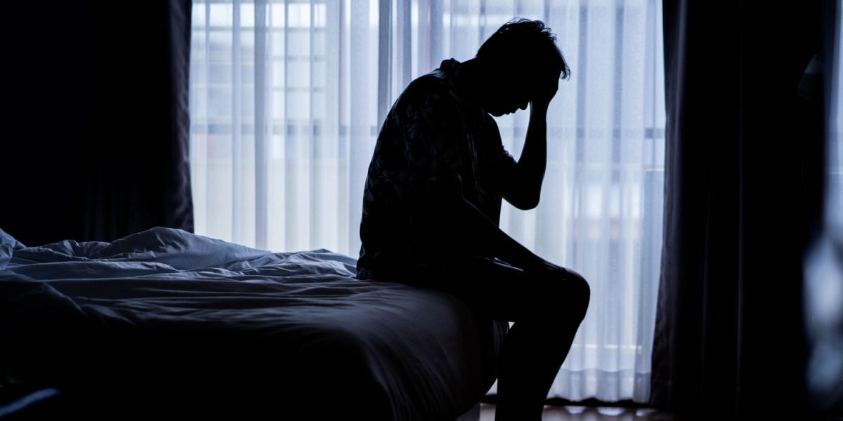 A dark silhouette of a man sitting at the edge of his bed and his hand supporting his head looking dejected