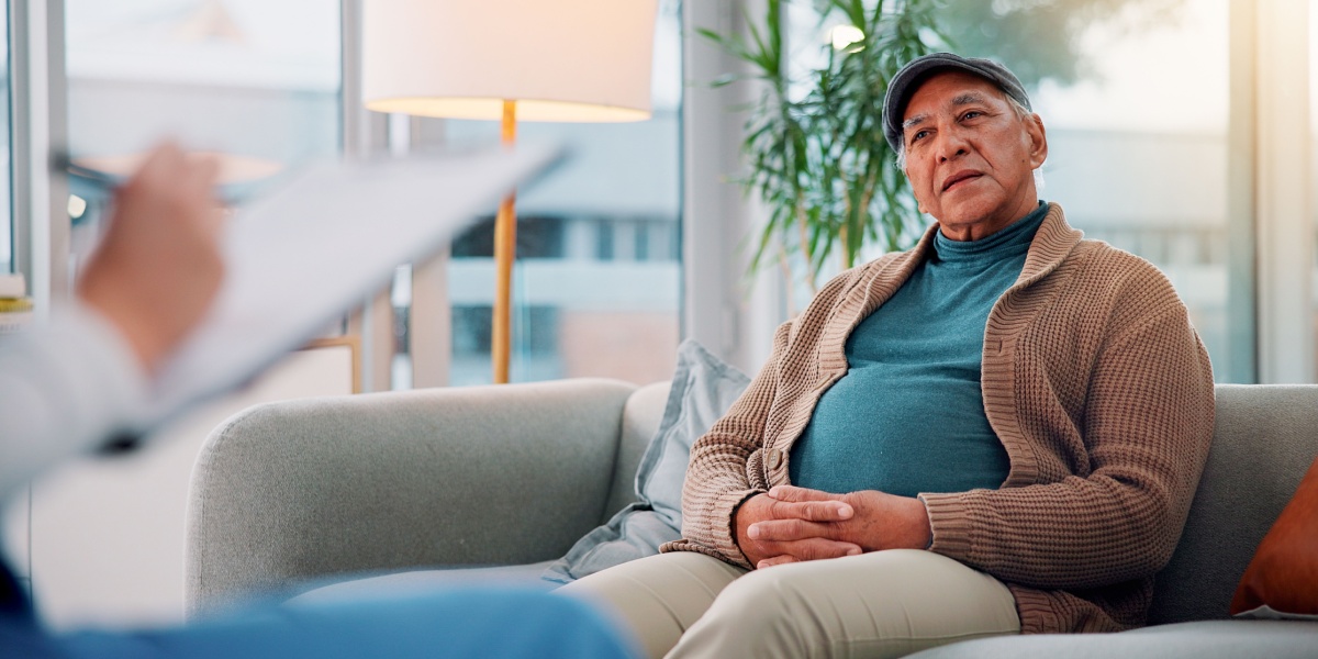 A bright photo of an elderly man sitting on a couch and a counselor in front of him.