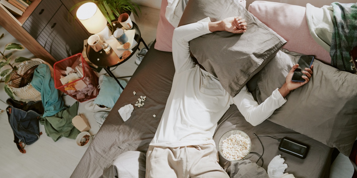 A bird's eye view of a messy room and a man in the middle of the bed covered with a pilllow over his head.